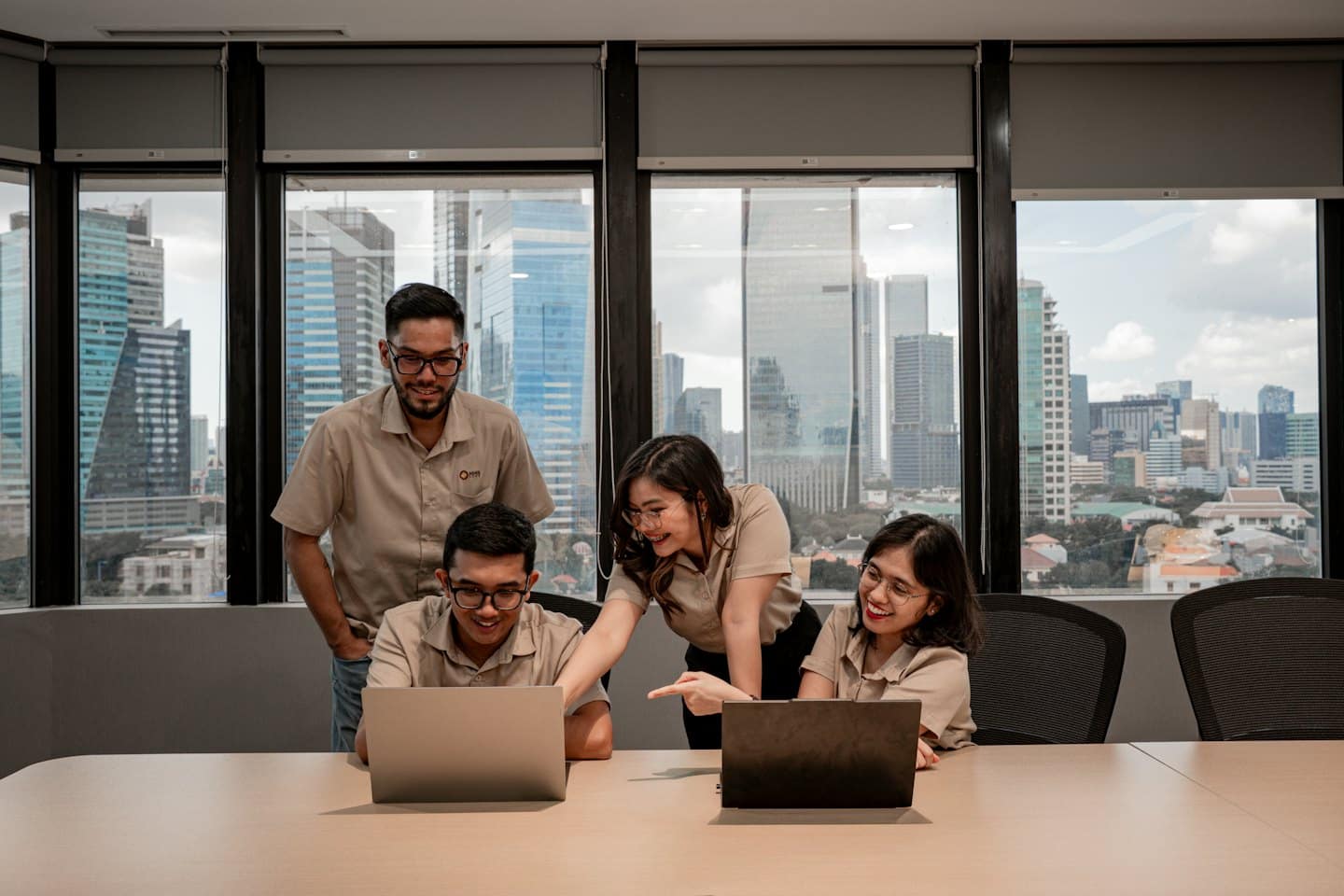 Four smiling professionals discussing work around laptops in an office.